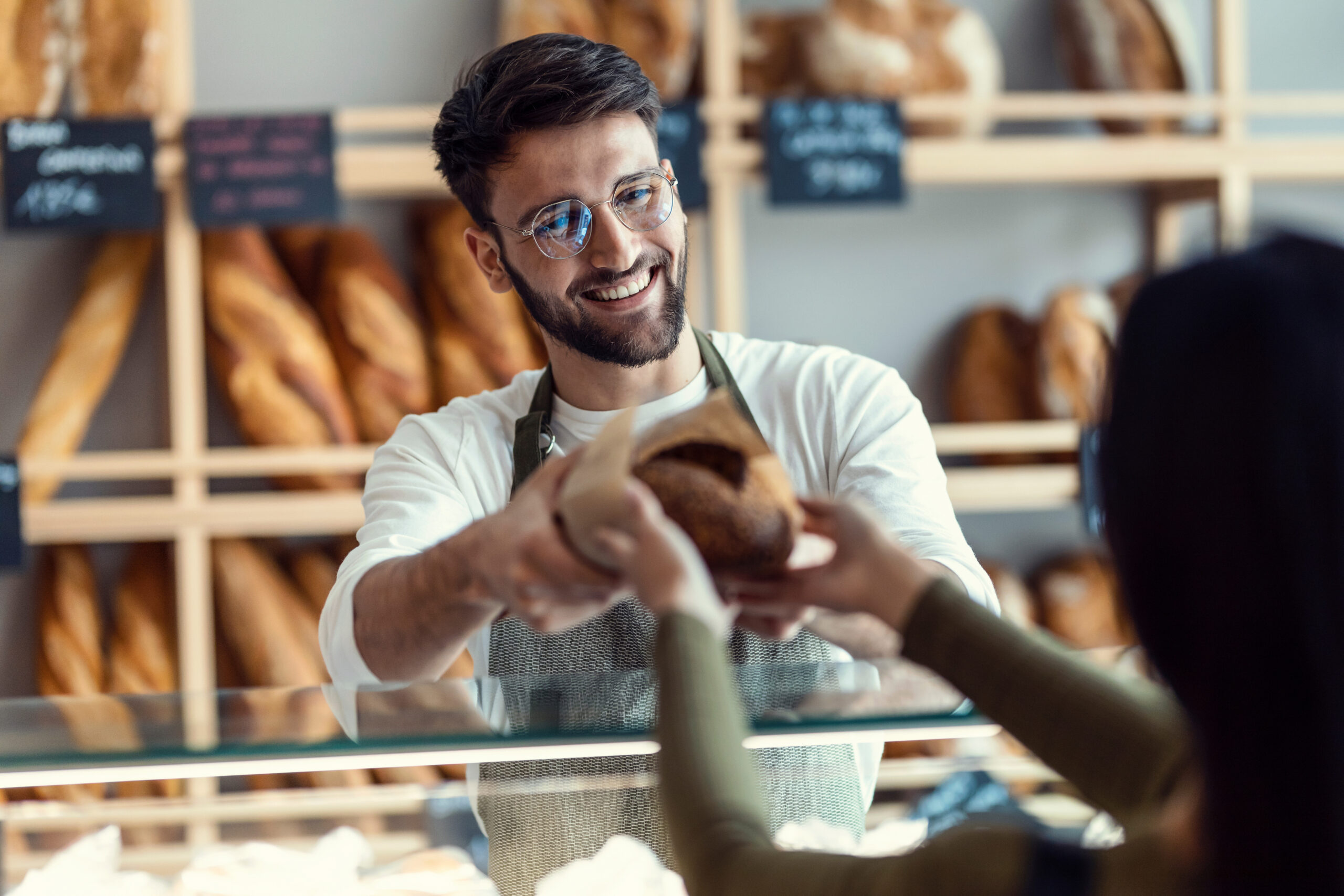 Shot of cheerful seller giving fresh loaf of bread to smiling woman in the pastry shop.